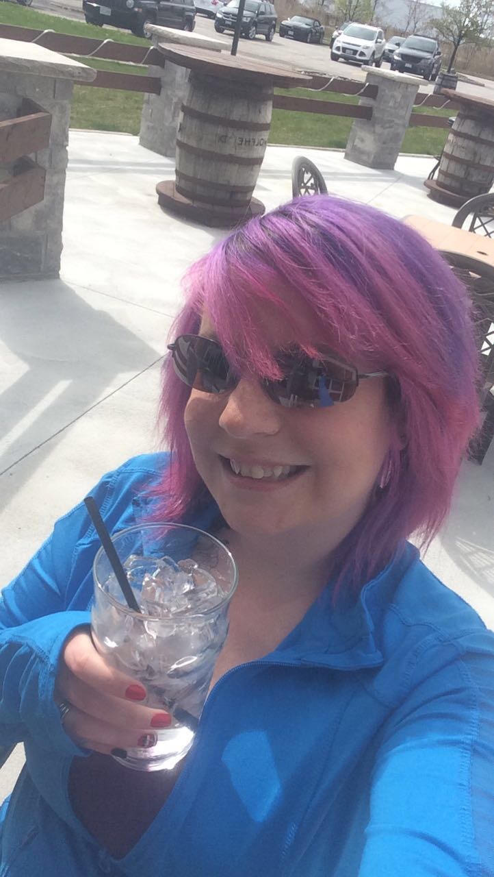 A woman smiling holding a drink and taking a selfie with pink and purple hair, sunglasses and a blue shirt