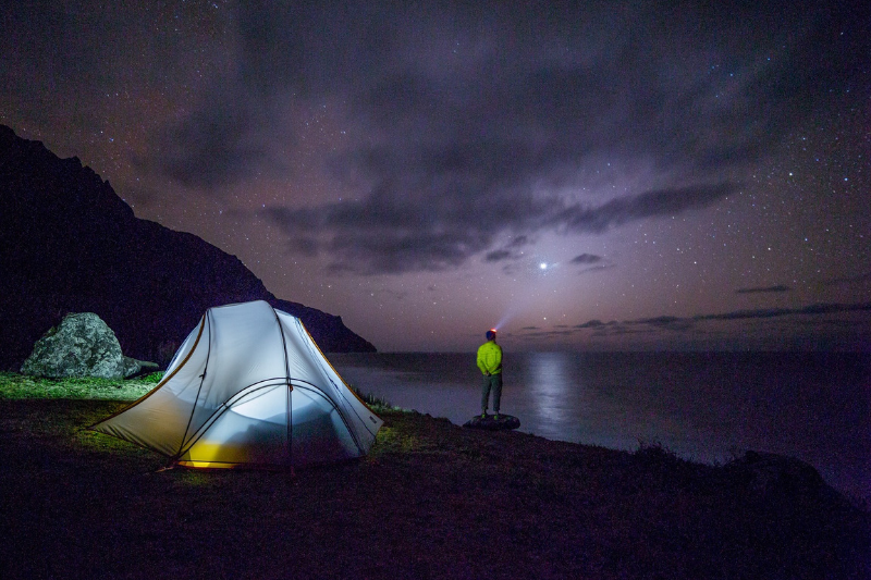 illuminated tent next to a body of water under the night sky with a lone camper standing on the shore