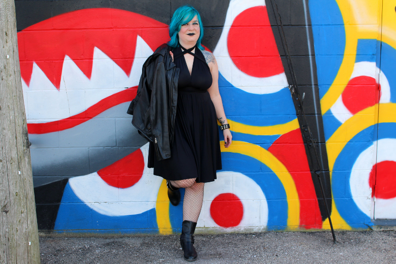 woman with blue hair in black dress, leaning against bright mural painted wall