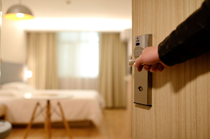 hand holding open a hotel room door, revealing a table and a bed
