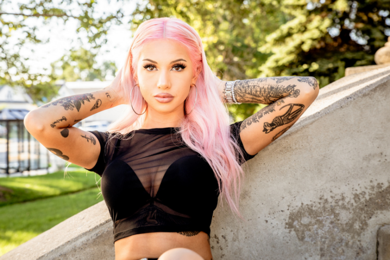 woman with tattoos and long pink hair leaning back against a cement wall, hands behind her head, in the sun