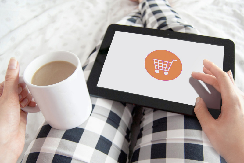 woman sitting in her grey plaid pajamas, holding a white mug of coffee and shopping online on her tablet