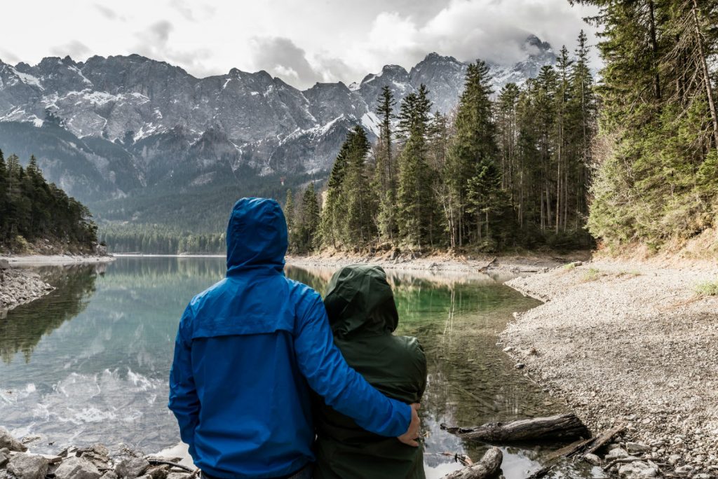 a couple standing together, one with their arm around the other, wearing rain coats and staring out across a lake with the mountains in the background