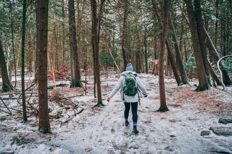 hiker in a white jacket with a green backpack, hiking through a wooded area in the snow