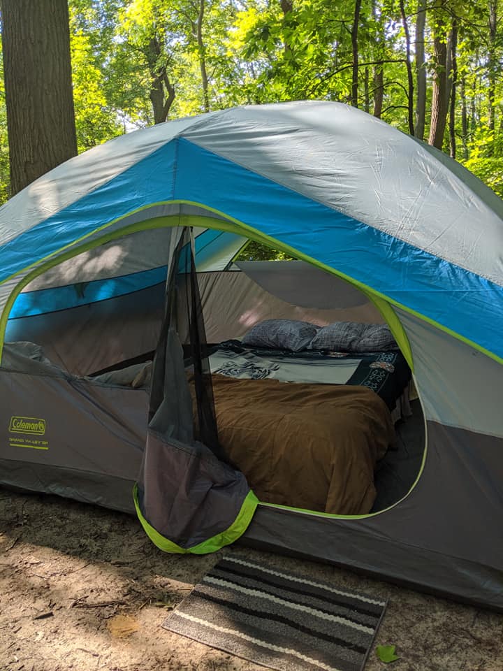 coleman tent set up in a wooded area with the door open, a bed inside
