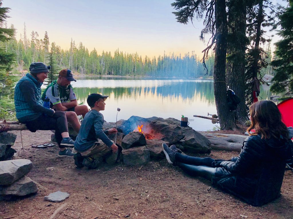 family gathered around a campfire pit beside a lake, roasting marshmallows