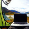 computer sitting on the end of a sleeping bag inside a green tent with the tent door open showing a number of other tents set up beside a lake