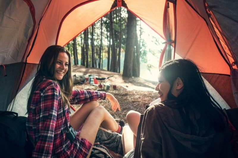 two brunette women sitting in an orange tent with the door open, showing a forested area with camping gear