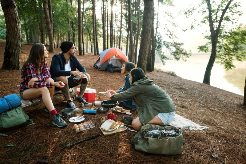 group of 4 people eating outside in a forested area with a tent in the distance