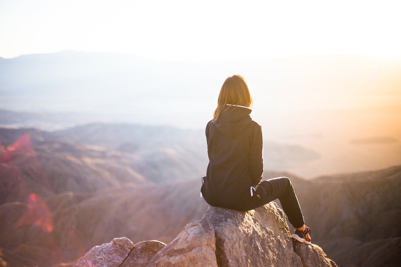 woman in a black hoodie, black pants and sandal sitting on a cliff edge, overlooking a foggy view