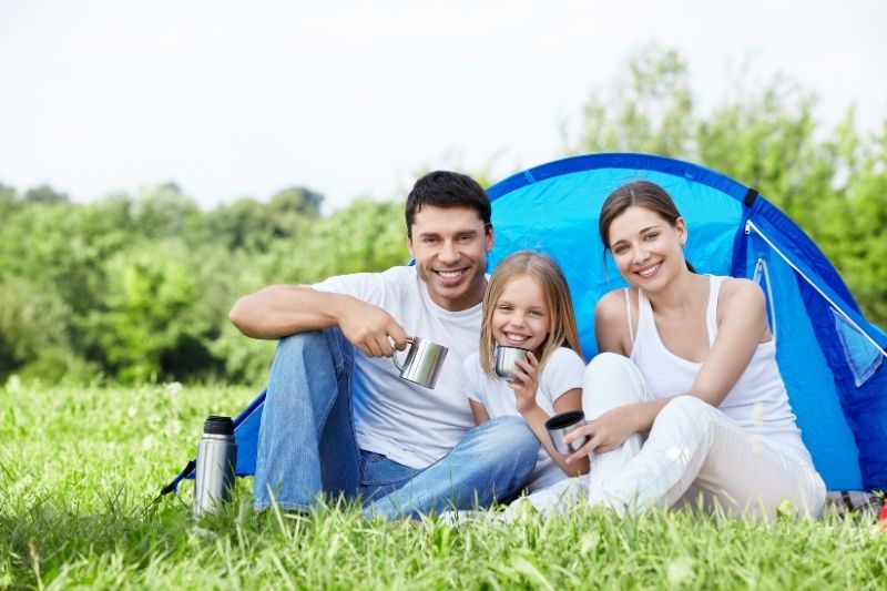 A small family including a man, woman and a young child are sitting in front of a blue tent outdoors. They are wearing white t-shirts and blue jeans (white for the child and woman) while holding stainless steel mugs. Next to the man is a stainless steel thermos. 